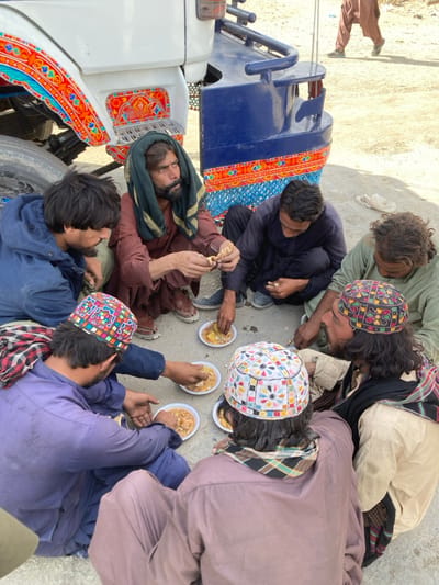 Seven adult males are observed sitting on a light-colored, dusty ground surface in an outdoor setting in Chaman City Tehsil, Pakistan. They are gathered in a loose circle, engaged in a communal meal. Six individuals are actively consuming food from small, disposable white plates placed on the ground, using their hands to eat a yellowish, stew-like substance, possibly with bread. Their attire consists of traditional garments, including shalwar kameez-style clothing in various colors such as brown, blue, green, and purple. Two men in the foreground wear distinctive, colorful embroidered caps. Partial views of a white vehicle, likely a truck or bus, with vibrant traditional Pakistani truck art in red, orange, and blue patterns, are visible in the background, along with a blue metal bumper. Another individual in brown attire is partially visible walking in the distant background.