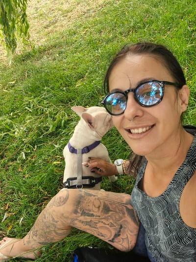 An adult female, with dark hair and glasses, is taking a selfie while sitting on grass next to a light-colored dog. The female is smiling and wearing a patterned tank top and has extensive tattoos on her legs. The dog is wearing a harness and is facing away from the camera. The scene appears to be outdoors, possibly in a park or grassy area. The location is not specified in Bridlington, United Kingdom.