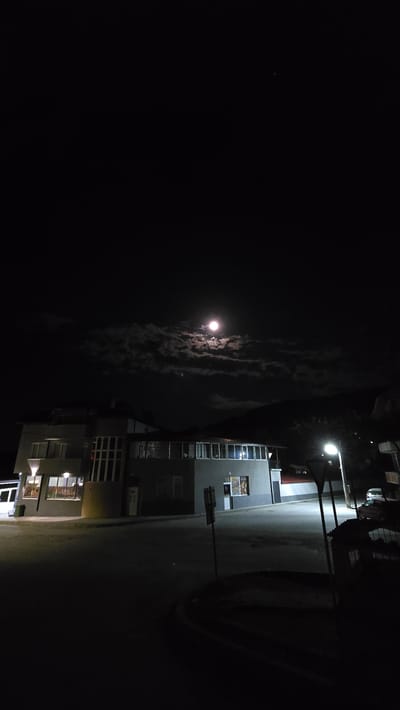 A nighttime scene in Breznitsa, Bulgaria, shows a street illuminated by a streetlight. A building stands next to the road, with several lit windows. A full moon shines brightly through the clouds. The sky is dark, with a few visible stars.
