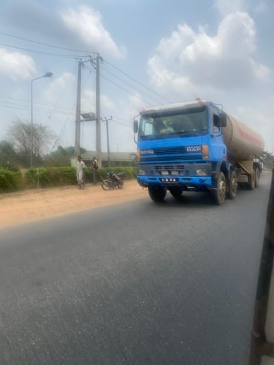 This image captures a roadside scene in Odo Ona, Nigeria, under a hazy daytime sky with scattered clouds. The primary subject is a blue DAF 85 CF tanker truck, identifiable by "DAF 85 CF" and "LEYLAND" branding on its cab, which pulls a light-colored, cylindrical tanker trailer. The truck is positioned on an asphalt road, occupying the right and central portions of the frame, facing slightly right from the viewer's perspective.
On the unpaved dirt shoulder to the left of the truck, two adult males are standing. One male wears light-colored clothing, while the other wears a light-colored vest over a darker garment with dark trousers. Adjacent to them, a dark-colored motorcycle is parked upright on its kickstand.
The environment includes a series of utility poles with multiple power lines and a visible transformer assembly running parallel to the road on the left. A single streetlight pole is also present further down the road. Beyond the shoulder, sparse vegetation, including trees and bushes, a fence, and distant, low-rise structures are visible. The asphalt road surface shows minor wear. The scene is brightly lit, indicative of daylight conditions.
