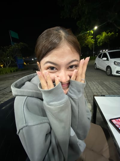 A young woman is captured in a medium shot at night, outdoors, with a street lamp and trees visible in the background. She is wearing a gray hooded sweatshirt and has her hands near her face, a gesture that could be interpreted as playful or shy. Her hair is brown and pulled back. A white car is parked behind her, and a white table with a phone on it is to her right. The scene is likely in Mekar Bakti, Indonesia, given the context.