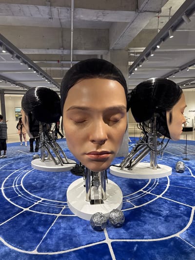 The image shows a contemporary art installation likely in a museum or gallery in the Nanshan District, China. Three large, hyper-realistic human head sculptures are prominently displayed on white circular plinths on a bright blue carpet with a geometric white line pattern. The heads are positioned at varying heights, appearing to be stacked vertically in the frame.

The faces of the sculptures have closed eyes. Under each head is a series of metal tubing that forms a mechanical neck and base.

In the background, people are observing the artwork. A row of track lighting fixtures is visible on the left side of the image. The overall environment has an industrial aesthetic with exposed concrete pillars and metal grates.