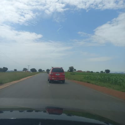 A red Ford Galaxy minivan is traveling on a paved road in Dorowa, Nigeria. The minivan is seen from behind, and it has a roof rack. The license plate is visible and reads "GLGT 590 JL". The road is bordered by a grassy embankment on the left and a field of crops on the right. In the distance, there are scattered houses and trees, and a range of mountains under a partly cloudy sky. The reflection of the minivan and the surrounding landscape can be seen on the dark surface of the vehicle from which the photo is taken, suggesting a driver's perspective.