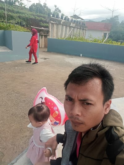 This image captures an outdoor scene in what appears to be Pangalengan, Indonesia, during daylight hours under an overcast sky.
In the foreground, a man is taking a selfie, looking directly at the camera with a serious expression. He wears a khaki or olive-green jacket over a grey hooded top and a pink inner shirt. To his left, a baby or toddler, wearing a light pink dress over a white long-sleeved top, is partially visible, facing away from the camera. Next to the baby is a pink, M-shaped balloon.
The subjects are positioned on a light-colored, paved surface, possibly a terrace or elevated area. In the middle ground, a woman dressed entirely in red, including a matching hijab, is walking from left to right. She wears black sneakers. Behind her, there are low blue-grey walls bordering the paved area.
The background reveals a hilly, green landscape typical of a rural or semi-rural area. A concrete wall topped with several potted plants runs across the mid-distance. Behind this wall, a house with a reddish-brown roof is visible. The sky is largely overcast, suggesting soft lighting. No discernible text is visible in the image.