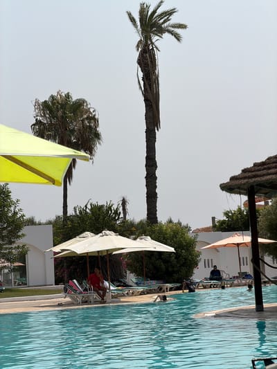 This image depicts a resort pool area in Sousse, Tunisia, on a bright, hazy day. The foreground is dominated by the clear turquoise water of the swimming pool, showing ripples and reflections. Along the edge of the pool, lounge chairs are arranged, some occupied by people. A man in a red shirt and shorts sits on a lounge chair under a large white umbrella. Several other umbrellas and lounge chairs are visible throughout the scene. Palm trees, both tall and short, are scattered around the resort grounds, with one particularly tall palm tree standing prominently in the center-right of the frame. Behind the pool area, white buildings with arched windows and doors are visible, suggesting hotel or resort accommodation. In the distance, part of a water slide can be seen. The overall impression is that of a relaxed holiday atmosphere.