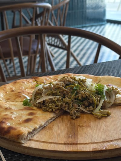 A close-up, eye-level shot shows a piece of flatbread topped with a mixture of what appears to be small seeds or grains, onions, and herbs, served on a wooden board. The flatbread is golden brown with some darker blistered spots. The filling is a light brown mixture with visible green herbs and white onion slices. The wooden board has a circular groove. In the background, out of focus, are wooden chairs with a patterned floor and a vertical glass pane. The scene is likely set in a restaurant in Gamarjveba, Georgia, given the context.