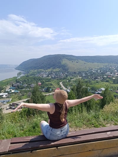 An aerial view captures a person sitting on a wooden bench, facing away from the camera, with arms outstretched. The person is wearing a brown tank top, denim shorts, and a straw hat. They are positioned on a grassy hillside overlooking a valley. The valley contains a village with houses, a winding road, and a large river to the left. The river is bordered by green reeds and a distant forested hill. The sky above is blue with scattered white clouds. The scene appears to be the city of Solnechnaya Polyana, Russia, specifically a viewpoint that offers a panoramic vista of the surrounding landscape and settlement.