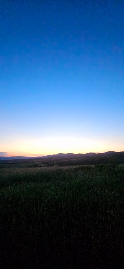 An outdoor landscape photograph captured at dusk shows a vast field of dark green grass under a deep blue sky that transitions to lighter hues near the horizon. Distant, undulating hills are silhouetted against the twilight sky. The image is devoid of any human presence, structures, or specific identifiable landmarks. It is a scene of natural, undeveloped terrain.
