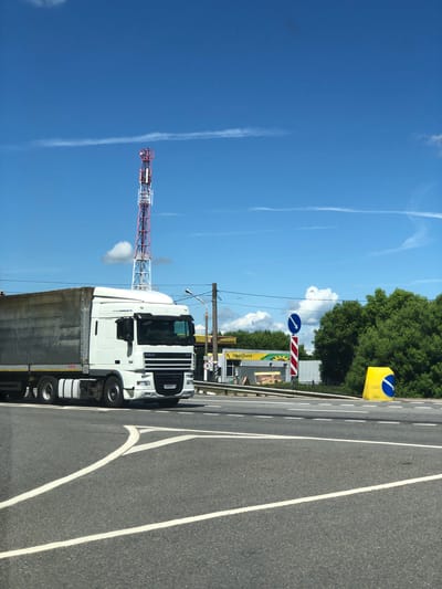 A white semi-trailer truck with a gray trailer drives on a road in Izdeshkovo, Russia, on a sunny day with a bright blue sky and scattered white clouds. The truck is in the foreground, occupying the left and central portions of the image. It has a "DAF XF" logo on the front. In the background, there is a red and white cell tower, a yellow and white gas station with "ROSNEFT" and "PODSOLNUH" signage, a variety of trees, and road signs. The road is paved with asphalt and marked with white lines, indicating multiple lanes and an intersection. There are no people visible in the image.