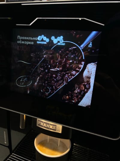 A coffee machine displays an image of roasted coffee beans in a scoop on its screen. Text in Cyrillic is visible, likely advertising the "correct" roasting of the coffee. Next to the screen, a cup of coffee is dispensing from the machine. The brand name "FRANKE A600" is prominently displayed on the machine's side. The overall scene suggests a modern, automated coffee-making process.