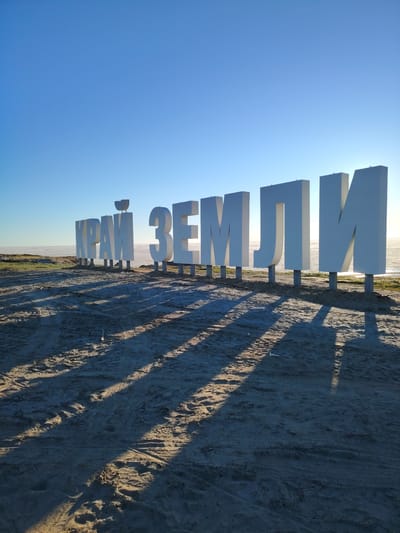 A bright, clear, sunny day shows large, white, three-dimensional block letters spelling out "КРАЙ ЗЕМЛИ" (Kray Zemli, meaning "Edge of the Earth") positioned in a horizontal line on a sandy plain. The letters are supported by individual grey pedestals and cast long, distinct shadows onto the sandy ground. In the background, a flat, expansive body of water, possibly a frozen sea or large lake, stretches to the horizon under a cloudless, pale blue sky. The area appears to be located in Yamburg, Russia, a remote city in the Yamalo-Nenets Autonomous Okrug known for its harsh Arctic climate. The scene is devoid of any people or animals. The environment is characterized by the vast, open, and sparsely vegetated landscape.