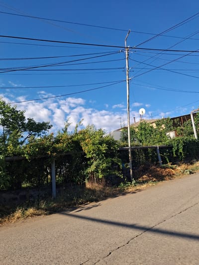 The image depicts a residential street in Yeghegnadzor, Armenia, bathed in bright sunlight under a clear blue sky dotted with white clouds. A weathered utility pole, adorned with numerous electrical wires, stands prominently on the right side of the frame, leaning slightly. Beyond the pole, a residential building with a red-tiled roof is partially visible, topped with a white satellite dish. Dense green foliage, likely trees and vines, obscures much of the property, forming a natural barrier along a stone wall that runs horizontally across the mid-ground. The foreground is dominated by a paved road with a visible crack running diagonally across it. The road surface appears somewhat aged. The overall scene suggests a typical, sun-drenched day in a quiet, possibly rural or suburban, area of Yeghegnadzor.