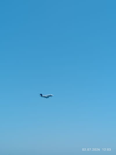 A white airplane with dark tail wings is flying in a clear blue sky. The plane is seen from below and is angled to the right. Its landing gear is extended, suggesting it is either taking off or preparing to land. The sky is a uniform bright blue, indicating fair weather and daylight.
In the bottom right corner of the image, white text displays a date and time: "02.07.2026 12:03". This suggests the photograph was taken on July 2, 2026, at 12:03 PM. The location context provided is Larnaca, Cyprus, which is known for its international airport, making the presence of an airplane a fitting detail for the setting. There are no people visible in the image, and the scene is dominated by the vast expanse of the sky and the solitary airplane.