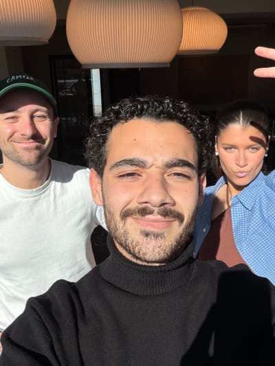 The scene is indoors in Marche-lez-Écaussinnes, Belgium, illuminated by strong natural light casting distinct shadows. Three individuals are positioned closely, appearing to be in a selfie photograph.

In the foreground, a male with dark, curly hair, a mustache, and a short beard smiles directly at the camera, wearing a black turtleneck sweater. His head occupies the central lower portion of the frame.

To his left and slightly behind him, a second male with short light-brown hair, wearing a green baseball cap with "CAMTAR" visible on the front, and a white t-shirt, also smiles at the camera.

To the right and slightly behind the foreground male, a female with dark hair pulled back from her face wears pearl earrings, a light blue plaid button-up shirt over a brown tank top. She puckers her lips in a "duck face" expression towards the camera. Her right hand is raised, forming a V-sign or peace gesture, with its shadow partially obscuring her right cheek.

The background is out of focus, revealing two large, ribbed, light-colored pendant lamps hanging from the ceiling. A dark interior space is visible behind the individuals, with a reflection of an outdoor scene or light-colored structure visible through a window-like aperture on the far left.