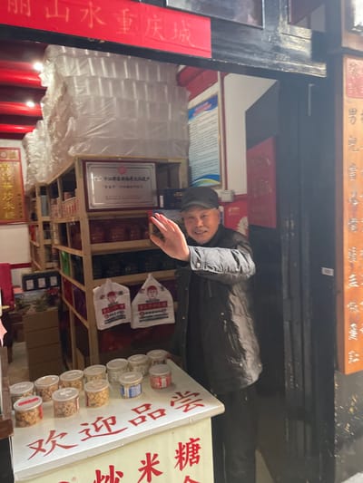 An elderly man wearing a black jacket and cap stands in front of a shop. Shelves behind him display bags and containers of goods. Signs with Chinese characters are visible. One sign reads "Ming Mountain Water Chongqing City". A certificate from the Jiangjin District Department of Culture is displayed. Another sign advertises "Changhua Fried Rice Candy" and "Welcome to taste". The shop appears to specialize in rice-based snacks and sweets.