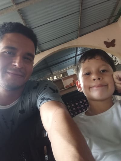 The image is a selfie taken by two people, an adult male and a young boy, possibly father and son. They are indoors, in what appears to be a residential setting with corrugated metal roofing and walls visible in the background. The lighting suggests it could be daytime, but the exact time is not discernible. The adult male is in the upper part of the frame, with his arm visible in the lower left. He is wearing a grey t-shirt with a black pattern. The young boy is in the lower part of the frame, leaning his head on his hand and smiling at the camera. He is wearing a white t-shirt. There is a brown butterfly decoration visible on a curved wall in the background, and some patterned fabric is also visible. The overall mood appears to be relaxed and happy.