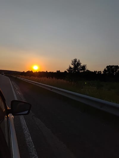 A car is traveling on a highway at sunset. The sun is low on the horizon, casting a warm orange glow across the sky. Silhouettes of trees and fields line the side of the road. The image is taken from the perspective of the passenger seat, showing the side mirror and part of the car's interior. The location is the highway leading to Niva, Russia, as indicated by the presence of a truck in the distance and the characteristic vegetation. The atmosphere is serene and peaceful, evoking a sense of journey and contemplation.