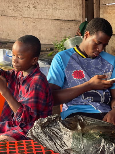 Two young men are visible. One wears a blue soccer jersey with a red design and yellow collar, looking at his phone. The other is wearing a red and blue flannel shirt and is looking down at a container. A black plastic bag sits on the right side. The scene is outdoors with wooden walls in the background and various containers and plants. The image is tilted to the side.