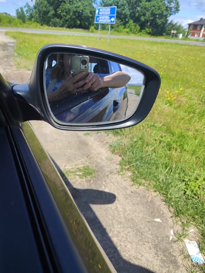 The image is a first-person perspective shot from inside a vehicle, looking into the side mirror. The mirror reflects a person holding a smartphone and taking a selfie. The background visible in the mirror and through the car window shows a roadside scene with green grass, trees, and a road sign. The road sign is blue with white text, indicating directions to "Matrosovo" and other locations. The environment appears to be rural or suburban, with a clear sky and daylight. The car appears to be stationary or moving slowly.