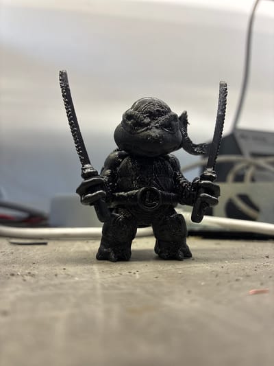 This is a close-up, vertically oriented photograph of a small, black 3D-printed figurine. The figurine depicts a stylized character, possibly a warrior or creature, with a rounded head and a body covered in textured lines indicative of the 3D printing process. It is holding two small swords, one in each hand, which also exhibit the layered texture of 3D printing. The figurine is positioned on a textured, light-colored surface, which appears to be a build plate or surface of a 3D printer. In the background, out of focus, are elements of a white and grey machine, likely the 3D printer itself, and a plain, bright background suggesting an indoor setting. There is no visible text in the image. The lighting is bright and seems to be coming from the side, creating some highlights and shadows on the figurine and the surface. The overall impression is of a recently printed object, showcasing the detail and texture achieved through 3D printing technology.