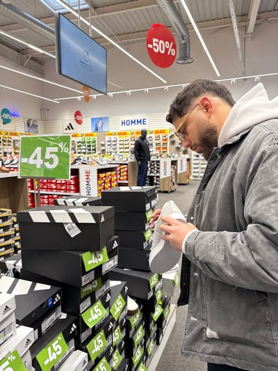 A male individual is depicted in the foreground within a retail shoe store in Reichstett, France, examining a white athletic shoe. He possesses short dark hair, a beard, and wears light-framed glasses, a light-colored hooded sweatshirt, and a dark grey, washed-out jacket. His head is tilted downwards, and he holds the shoe in both hands, visually inspecting its sole and upper surface. Adjacent to him and extending into the mid-ground are multiple stacks of black shoeboxes, identifiable as Adidas brand by their distinctive three-stripe design and visible logos. Numerous green square signs displaying "-45%" in white are affixed to these shoeboxes, indicating a discount.

The background reveals a typical retail interior with white walls featuring horizontal blue and white stripes. Extensive shelving units display an array of shoes, accompanied by additional stacks of shoeboxes. Overhead, circular red signs with "-50%", orange signs with "-40%", and a blue sign with "-60%" are suspended, alongside a large rectangular digital screen showing a loading or update message. Exposed silver air ducts and fluorescent lighting fixtures are visible on the ceiling. Further back, another male individual, wearing a dark jacket and cap, stands near a shoe display. A dark mannequin is positioned next to a "HOMME" (Men) sign, which is also flanked by "SOLDES" (Sales) banners and an Adidas logo.