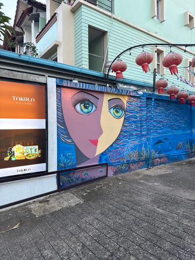 The image captures a vibrant street scene in Ban Bang Niang, Thailand. The focal point is a large mural painted on a textured wall. The mural depicts a stylized human face, split down the middle, with one half rendered in shades of purple and brown, and the other in yellow. Both halves feature striking, colorful eyes. To the right of the face, the mural transitions into an underwater scene, complete with coral reefs, fish, dolphins, and a turtle, all rendered in various shades of blue and white. Above the mural, red Chinese lanterns are suspended from a curved black wire, adding a festive touch. To the left of the mural is a large advertisement for "Takola Village," featuring images of clothing, accessories, and what appears to be a beverage or food product. The advertisement has an orange and white background. The street itself is paved with textured gray concrete, with visible lines and some patchy wear. The background shows elements of building architecture, including light blue and pale green facades with window frames. The overall impression is a colorful and lively urban environment.