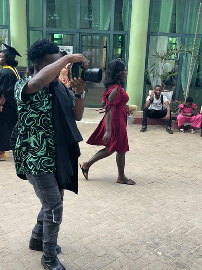 This image depicts an outdoor event scene in Madina, Ghana, on a paved area in front of a modern building with green-tinted glass windows and light green exterior elements.
In the immediate foreground, a male with dark, curly hair, wearing a black and mint green swirled patterned short-sleeve shirt, dark ripped jeans, and black boots, is captured in motion, holding a black and green professional camera with a long lens to his eye, focused on taking a photograph. A black garment, potentially a graduation gown, is draped over his left shoulder. He wears a watch on his left wrist and rings on his right hand.
In the mid-ground, a female with dark hair, wearing a maroon, knee-length dress with cut-out shoulders and ties on the sleeves, walks from left to right, carrying a woven basket on her right arm. She wears black flat sandals with gold accents.
In the background, to the far left, a person in a black graduation cap is partially visible. Further right, closer to the building, a male in a white t-shirt with a black star emblem (suggesting the Ghana flag) and black pants is seated on a low concrete ledge, holding a mobile phone to his ear. A black backpack is on his back. Adjacent to him, a female in a pink top with ruffled sleeves and pink pants is seated, looking down at a mobile phone in her hands. The ground throughout the visible area is composed of light-colored paver stones laid in a herringbone pattern. Potted plants are situated near the building's facade.