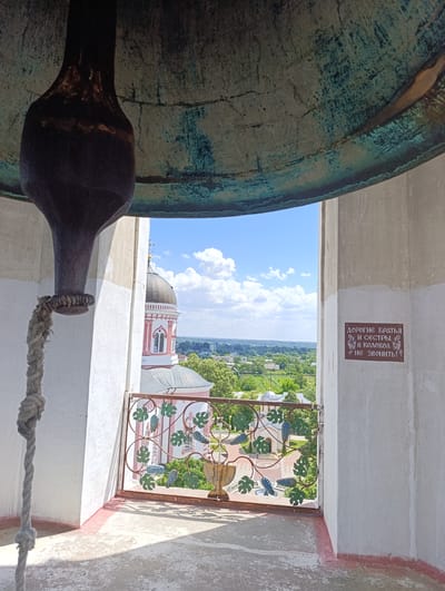 The image captures a view from the belfry of the Ascension Monastery in Kitskany, Moldova. In the foreground, a large bell is partially visible, with a thick rope hanging down. To the right, on a concrete wall, a sign in Cyrillic script reads "Дорогие братья и сестры, в колокол не звонить!" which translates to "Dear brothers and sisters, do not ring the bell!". Through the open archway of the belfry, a scenic view unfolds, showcasing the monastery's pink and white church with its dome and cross, surrounded by lush green trees and the landscape of the surrounding area. The sky is a clear blue with scattered white clouds. A decorative metal railing with a grape vine motif separates the belfry from the exterior view. The ground inside the belfry is a rough concrete surface.