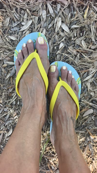 A close-up overhead view captures a person's feet wearing blue and green patterned flip-flops with bright yellow straps. The toes are adorned with metallic yellow nail polish. The feet are positioned on a bed of dry leaves and straw, suggesting an outdoor setting. The lighting appears to be natural daylight, casting subtle shadows. A partial view of the person's legs is visible, with a tattoo on the left ankle. The overall scene evokes a relaxed, summery atmosphere, possibly in a warm climate like Kuşadası, Turkey, given the context. There is no visible text in the image.