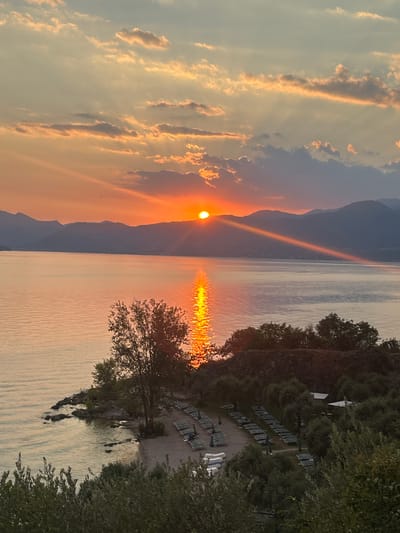 The image depicts a serene sunset over a lake, likely Lake Garda, Italy, given the context. The sun, a bright orb, is partially obscured by silhouetted mountains in the distance, casting a warm orange glow across the sky. Rays of sunlight fan out from the sun, illuminating scattered clouds with hues of orange and yellow. A shimmering reflection of the sun stretches across the calm, vast expanse of the lake.
In the foreground, a small, rocky shoreline meets the water. A sandy beach area is visible with rows of empty lounge chairs and umbrellas, suggesting a resort or recreational area. Several trees, some bare and others with dense foliage, are scattered across the land. A few small boats are pulled up on the shore. Lush green vegetation frames the bottom of the image. The overall impression is one of tranquility and natural beauty at dusk.