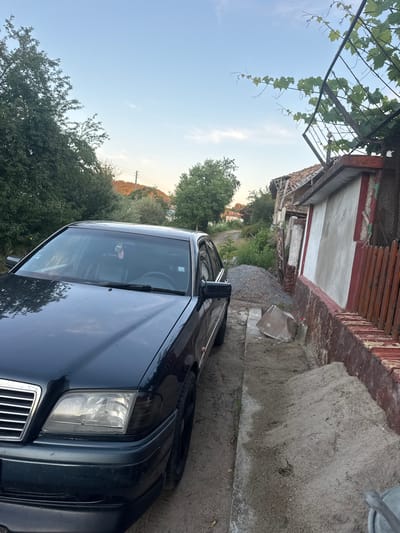 The image depicts a dark-colored sedan parked on a dirt pathway. To the right of the car is a red brick wall with a rough concrete and plaster facade above it. A wooden fence runs alongside the brick wall. Further to the right, a dilapidated building with a partially collapsed roof is visible. Above the building and the wall, a vine-covered trellis extends outwards. The background features lush green trees and a clear sky with a gradient of light blue to lighter hues near the horizon, where hills are faintly visible. The overall scene suggests an outdoor, possibly rural or semi-urban, setting. There are no people visible in the image. The environment appears to be in a state of disrepair, with loose gravel and sand on the ground. The location is identified as Dragoevo, Bulgaria.
