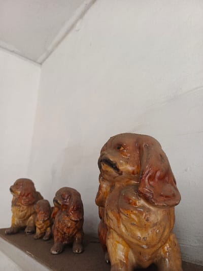 Four ceramic dog figurines of varying sizes sit on a shelf against a white wall. The dogs are brown and tan, with long ears and a seated pose. The largest dog is positioned prominently in the foreground, while the others are smaller and arranged in a line behind it. The shelf appears to be made of a dark material.