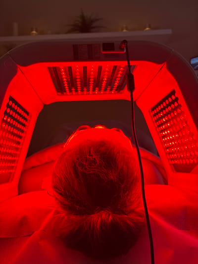 In this overhead, close-up shot, a person is undergoing a red light therapy treatment. The person is lying down, with their head tilted upwards, and their face is partially obscured by the red light-emitting device. The device itself is a large, horseshoe-shaped apparatus with numerous red LED lights arranged in panels. The intense red light bathes the person's face and hair, creating a warm, saturated glow.
The scene appears to be an indoor setting, likely a spa or treatment room, given the nature of the activity. The background is softly lit and out of focus, with hints of a white wall and possibly some plant foliage on the left side, suggesting a professional or domestic spa environment. The lighting is dominated by the red light therapy machine, casting a strong red hue over everything within its proximity.
The person's hair is dark and appears to be styled. The skin on their face is illuminated by the red light. The overall impression is one of a therapeutic or cosmetic treatment, emphasizing relaxation and rejuvenation through light therapy. There is a visible label on the side of the device with text in English, though it is too small to be legible. The time of day and weather are not discernible from the image. The emotions of the subject are not directly conveyed, but the context suggests a calm and possibly serene experience.