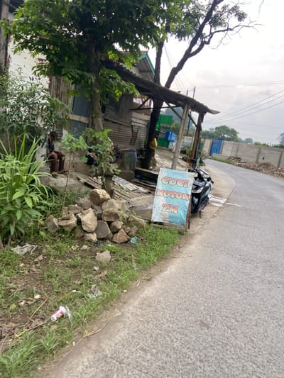 The image shows a motorcycle parked on the side of a paved road in front of a small building. The building is partially obscured by a large tree and a wooden awning. A person is visible sitting on a concrete step, wearing a blue shirt and black pants. A pile of rocks sits next to the person and motorcycle, indicating a possible construction site. A blue and white sign with the words "Cuci Motor ZM" stands beside the motorcycle. The sign is likely for a nearby motorcycle wash. The road is empty and the sky is overcast with a few white clouds. The scene is located on the side of a small, residential road in the Kecamatan Margaasih district, Indonesia, with the GPS coordinates of latitude: -6.964, longitude: 107.539. The image was taken from a perspective slightly above ground level. There is a single empty plastic cup laying on the ground.
