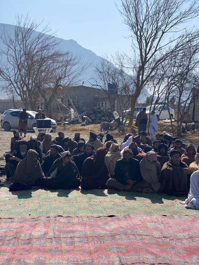 A group of people, primarily men, sit on a patterned mat outdoors. Most are dressed in traditional clothing with head coverings. Behind them are bare trees and a mountain range in the background. Several vehicles, including cars and vans, are parked nearby. A stone building is also visible in the distance. The scene is set under a bright, clear sky.