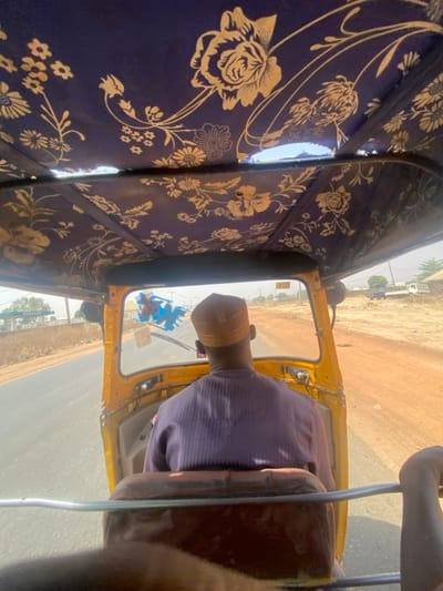 The image captures the interior of a yellow three-wheeled vehicle, likely a motorized rickshaw, in Baganakwo, Nigeria, viewed from the perspective of a rear passenger looking forward. The vehicle's roof is lined with dark blue fabric adorned with a repeating pattern of light-colored floral and foliage motifs; a small tear is visible in the fabric on the upper right side.
A single male individual is seated in the driver's position, viewed from behind. He wears a light purple, possibly striped, long-sleeved garment and a light brown, mesh-textured kufi-style cap. Blue plastic decorative items hang from the rearview mirror, visible through the windshield.
The vehicle is in motion on a paved asphalt road, which appears to be a two-lane thoroughfare. The roadside consists of dry, reddish-brown earth with sparse, desiccated vegetation. In the distance on the left, a structure featuring a green and white canopy is visible, consistent with a fuel station. On the right side, further from the road, indistinct buildings and a white truck are discernible against a clear, bright sky.