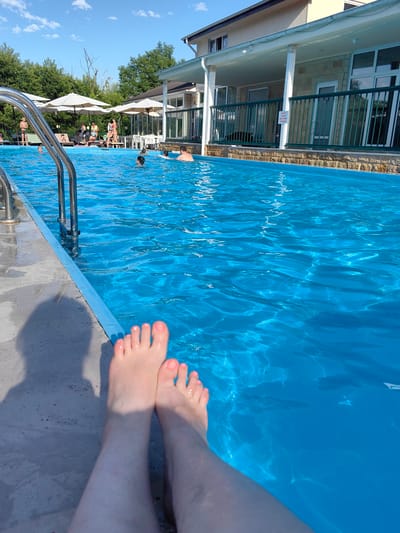 This is a medium shot, eye-level, outdoor photograph taken at a swimming pool, likely at the Oryol-Izumrud resort in Oryol, Russia, during daytime. In the foreground, two bare feet with pink toenail polish are resting on a grey concrete pool deck, partially submerged in the bright blue water. The feet cast a shadow on the deck. In the midground, the pool is filled with people swimming and relaxing. Several individuals are visible in the water, including children and adults. Along the edge of the pool, there are lounge chairs and white umbrellas providing shade. In the background, a multi-story building with a covered patio area stands adjacent to the pool. The building has light-colored siding and a tan stone foundation. Green metal railings line the patio. Lush green trees and a clear blue sky with scattered white clouds complete the environment. The overall scene depicts a leisure activity at a resort.