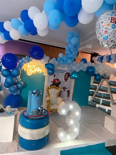 The scene is an indoor birthday party setup in Bonadikombo, Cameroon, located at latitude 4.048 and longitude 9.218. A blue and white balloon arch decorates the ceiling. An illuminated "Happy Birthday" sign hangs on a wall with a blue and turquoise backdrop. A white, illuminated "3" sits on the floor. A white throne chair is positioned to the left of the "3," and behind it is a photograph of a young girl. A blue and gold layered cake sits on a stack of three tires behind the "3." A small white table sits to the left of the cake, with a stack of white boxes on top. There is a small, white, square platform to the right of the number "3" with a few small white boxes on top. To the right of the throne, a white, wooden ladder leans against a wall decorated with a blue backdrop. There are small, blue boxes and packages stacked on the wall. The backdrop behind the ladder is white. The scene appears to be in a large, indoor space.