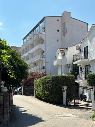 This media file captures a street scene in Lapčići, Montenegro. The image is oriented vertically, presenting a view of multi-story buildings with balconies and air conditioning units. The architecture appears to be of a residential or hotel type, with white facades and some decorative elements. A tall, manicured hedge lines the right side of the scene, bordering a paved walkway or road. A black wrought-iron gate is visible, with a motorcycle parked behind it. Further along the road, a silver car is partially visible.

The foreground features lush green foliage, including trees and vines, adding a natural element to the urban setting. A shadow of a tree is cast across the paved surface, indicating it is daytime. The sky is a clear, bright blue, suggesting fair weather. The overall mood is serene and quiet.

There is some text visible on a sign at the bottom right of the image, which appears to be in a Slavic language. One part of the text reads "MACA AKLIJA" and "20-E". The scene suggests a sunny day, likely during warmer months given the greenery. The location context of Lapčići, Montenegro, places this scene within a Mediterranean or Balkan environment.
