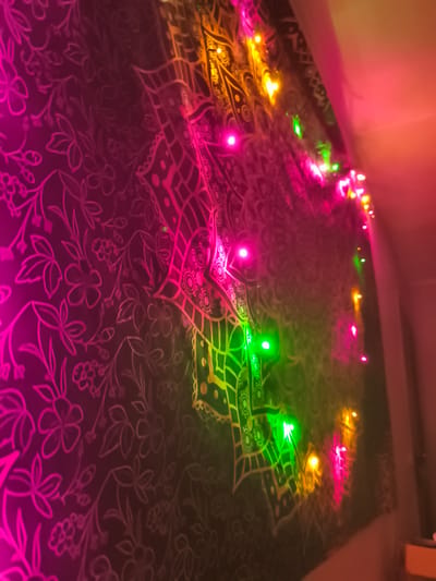 The image depicts a wall tapestry with a floral and mandala pattern, illuminated by colorful fairy lights. The lights cast pink, green, and yellow hues onto the tapestry, creating a vibrant and somewhat blurry effect. The tapestry appears to be dark in color with lighter outlines of the patterns. The scene is dimly lit overall, with the lights being the primary source of illumination. There is no indication of the city of Ekibastuz, Kazakhstan, in the image.