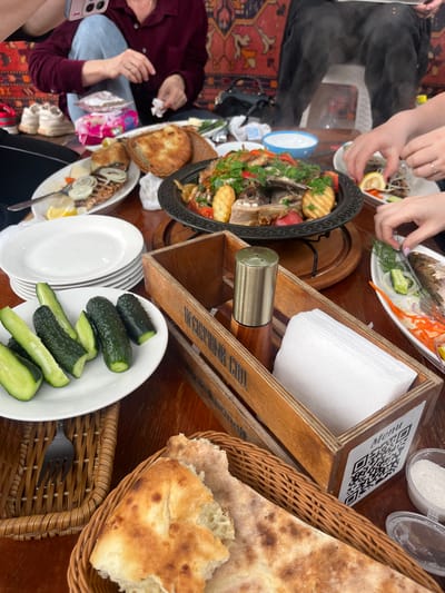 The image captures a high-angle, close-up view of a shared meal on a wooden table. Several people are seated around the table, engaged in eating and socializing. Food items include a large platter of meat and potatoes, a dish of fish, bread in a wicker basket, and a plate of sliced cucumbers. Utensils, plates, and a wooden napkin holder are visible. The QR code on the napkin holder suggests an interactive menu. The table is set on a rug. The photograph appears to be taken indoors. Based on the "Menu" visible on the napkin holder, the location may be Miatli, Russia.
