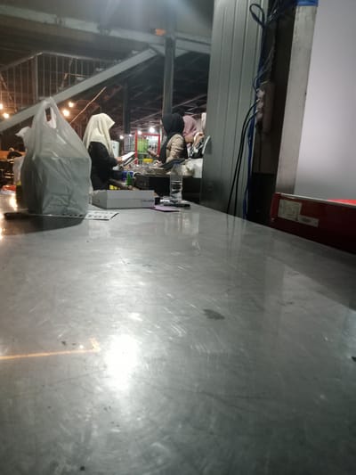 The image depicts an indoor scene, likely a food stall or market in Sukomoro, Indonesia. A stainless steel countertop dominates the foreground, reflecting overhead lighting. Behind the counter, two individuals wearing hijabs appear to be working or interacting with customers. One individual is wearing a white hijab and a black garment, while the other is wearing a dark hijab and a beige jacket. On the counter, there is a white plastic bag, a box labeled "REDMI Pad 2", a blister pack of pills, a glass of water, and other miscellaneous items. Electrical wires and a gray metal wall are visible to the right of the frame. The setting suggests a casual, possibly late-evening, atmosphere.