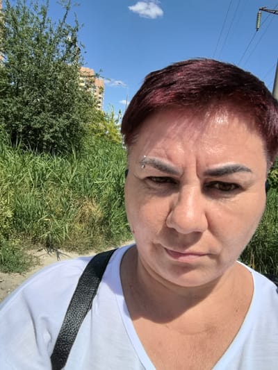 An outdoor, waist-up, selfie-style photograph captures a woman with short, burgundy-colored hair, an eyebrow piercing, and a white t-shirt. She is looking directly at the camera with a neutral expression. The background includes a tall, green grassy area, a building, and a bright blue sky with a single white cloud. The scene appears to be set in Verkhnetemernitsky, Russia.