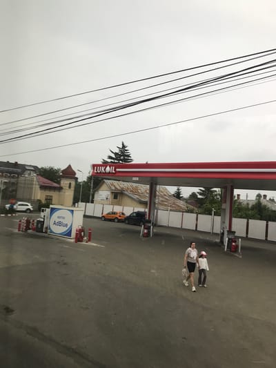 This image captures a wide-angle view of a Lukoil gas station in Roman, Romania, under an overcast sky. The prominent red and white Lukoil sign and canopy dominate the right side of the frame. Below the canopy, multiple fuel pumps are visible, along with red fire extinguishers and red-and-white striped bollards. A standalone blue and white dispenser unit for "ADITIVI AdBlue" is positioned closer to the foreground on the left.
Two individuals, an adult female and a child, are walking from left to right across the paved asphalt forecourt in the lower-middle portion of the image. The adult female wears a light-colored top, dark shorts, and white athletic shoes, carrying a light-colored bag in her left hand. The child, wearing a white long-sleeved top, grey pants, white athletic shoes, and a pink baseball cap, walks slightly behind and to the right of the adult.
In the background, beyond a white fence, various buildings are visible. These include residential structures and a building with a distinctive reddish-brown, multi-tiered dome or spire on the left, suggesting a church or historical building. An orange car and a dark SUV are parked near the gas station entrance, while a white SUV is visible further left next to a red "STOP" sign. Numerous black utility wires crisscross the grey sky in the upper half of the image. The overall environment appears functional and suburban.