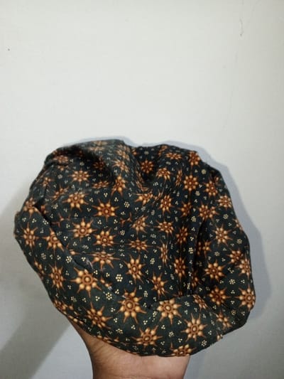 A dark-skinned hand, likely belonging to an adult, is depicted holding a crumpled fabric object against a plain, light-colored background. The fabric object appears to be a hat or a folded piece of cloth, characterized by a dark base color, possibly black or dark blue, adorned with a repeating pattern. This pattern consists of orange or reddish-brown star-like or floral motifs with radiating points, interspersed with clusters of smaller white or light-colored dots. The object is positioned centrally in the frame, casting a faint shadow on the background wall. The setting appears to be indoors, indicated by the uniform wall, with no specific environmental features visible that would identify the location as Na Sang, Laos.