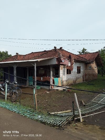 A view of a damaged brick house with a tiled roof, located in Kecamatan Anjatan, Indonesia, with coordinates of -6.389 latitude and 107.927 longitude. The house has a partially collapsed roof and a broken gate in front. There are two motorcycles parked in front of the house. The ground is covered in dirt and grass. Wires and cables are running across the sky.