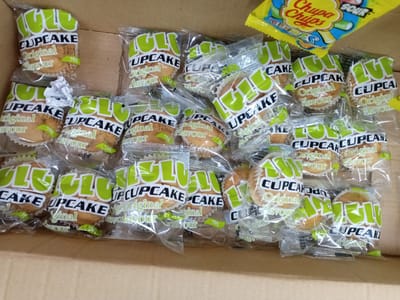 The scene, located in the city of Kawaji, Nigeria, depicts numerous individually packaged "LULU CUPCAKE Original Flavour" items within a cardboard box, alongside one packet of "Chupa Chups SOUR" candy.
Approximately 25 to 30 "LULU" branded cupcakes are visible, each enclosed in clear plastic packaging. The packaging prominently features "LULU" in large, light green lettering with a white outline, "CUPCAKE" in white capital letters, and "Original Flavour" in a cursive, yellowish-brown script. A green rectangle on each package indicates "70g." Several packages show date markings, such as "020624 / 280726" or "030424 / 280726," suggesting production and expiration dates. The cupcakes are light brown, consistent with a baked good, and are set in white fluted paper liners.
The items are loosely arranged inside a brown cardboard box, which constitutes the immediate environment. Some cupcake packages are stacked, while others lie individually across the box's base.
In the upper right portion of the box, a yellow packet of "Chupa Chups SOUR" candy is visible. The packet displays the "Chupa Chups" logo in red script, "SOUR" in yellow and blue, and an illustration of green and yellow stick candies.
No individuals are present in the image. The lighting suggests an indoor setting.