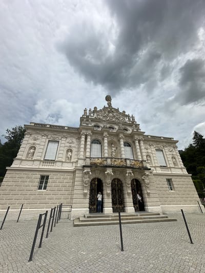 This is a ground-level, wide-angle shot of the facade of Linderhof Palace, located in Ettal, Germany. The palace is a grand, white Baroque-style building with ornate decorations, including sculptures and intricate carvings. The main entrance features large, gilded double doors and a balcony above. The sky is overcast with dramatic, dark clouds. In front of the palace is a cobblestone courtyard with a low metal barrier and posts. Two people are visible near the entrance, one entering and one standing by the doorway. Trees and foliage are present on the sides of the building, suggesting it is surrounded by a park or gardens. The overall atmosphere is imposing and historic.