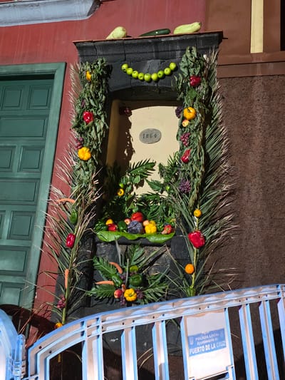The image shows a decorative display of fruits and vegetables adorning a doorway. The display is elaborate, featuring a variety of colorful produce such as peppers, grapes, apples, carrots, and what appear to be gourds. The doorway itself has a plaque with the number "1864" on it. The building has a red facade and a green door. In the foreground, there is a blue metal railing with a sign that reads "POLICIA LOCAL Ayuntamiento de PUERTO DE LA CRUZ," indicating the location as Puerto de la Cruz, Spain. The lighting suggests it is nighttime or dusk. There are no people visible in the image.