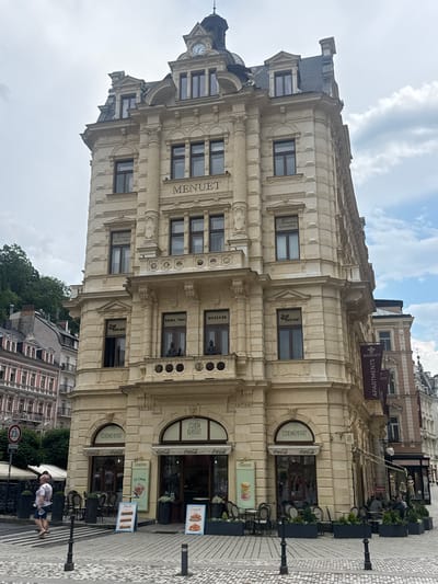 A tall, ornate, light-beige building with multiple stories dominates the frame, set against a sky with scattered clouds. The architecture features classical elements such as columns, detailed carvings, and a mansard roof topped with a cupola. On the facade, the word "MENUET" is prominently displayed in large, dark letters above a row of windows. Below this, a balcony runs across the building's face, supported by decorative brackets. Various storefronts occupy the ground level, including one labeled "MAD RABBIT" with "Coca-Cola" awnings, and another for "ICE CREAM." In front of these establishments, outdoor seating with chairs and tables is arranged.

To the left of the main building, a street sign with a red circle and "23t" is visible, indicating a weight limit. A person wearing a patterned shirt and blue shorts is walking on the cobblestone sidewalk in the foreground. Further left, other buildings with distinct architectural styles, including red-tiled roofs and arched windows, are partially visible. To the right of the main building, a banner with "MENUET APARTMENTS" is hung. The scene appears to be a street in the city of Karlovy Vary, Czechia.